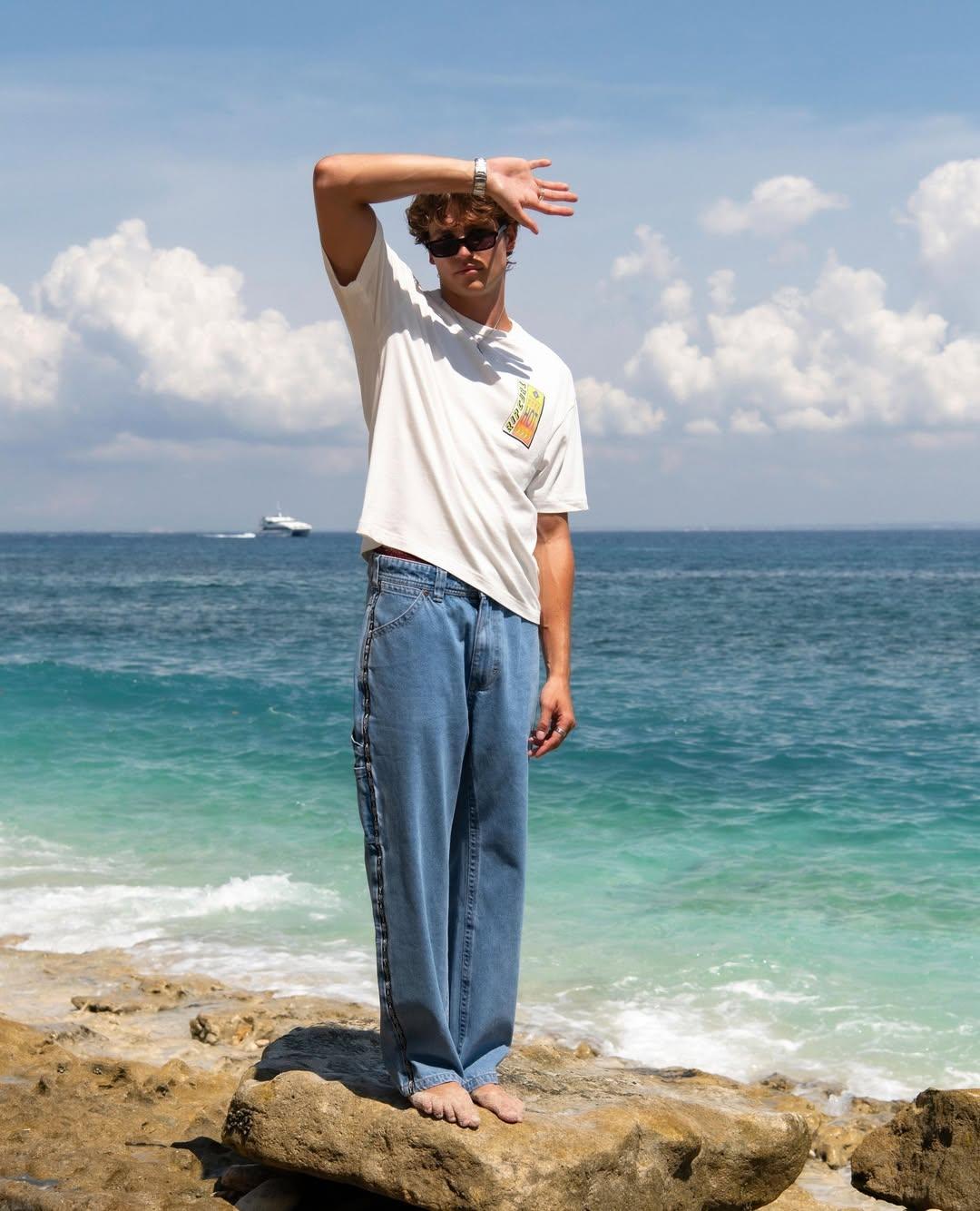 Person standing on a rock by the ocean with a white t-shirt and blue jeans.
